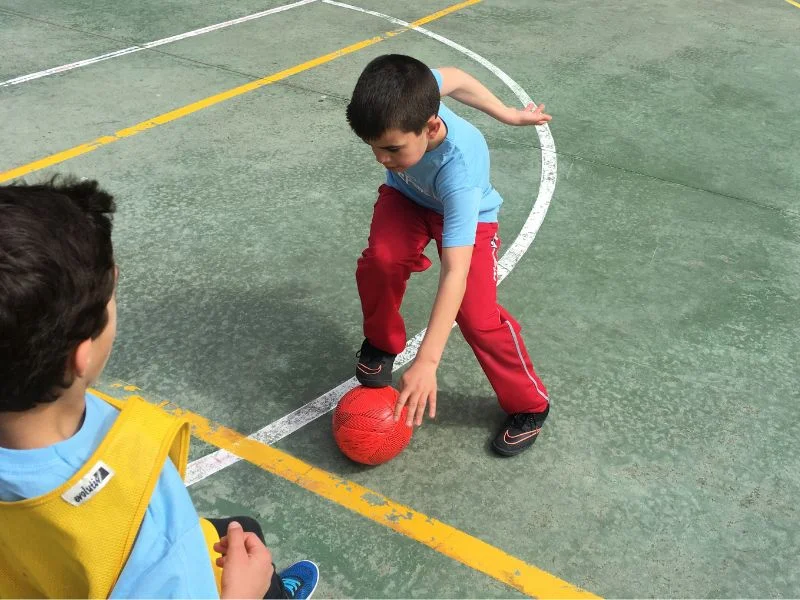 Dos nens jugant a bàsquet en una pista exterior durant una activitat extraescolar de futbol, mostrant habilitats esportives i treball en equip.